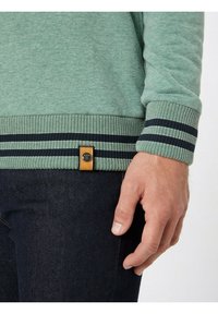Sudadera verde con ribete de rayas marino en el dobladillo, con una etiqueta de cuero marrón claro. Mano descansando sobre pantalones de mezclilla oscuros. Diseño y textura casual.