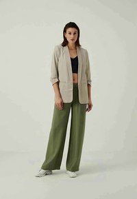 Femme debout, portant un blazer beige, un crop top noir, un pantalon large olive, des baskets blanches, avec des cheveux courts et foncés, sur un fond neutre.