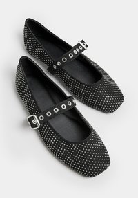 Zapatos planos de ante negros con punta cuadrada, adornados con tachuelas plateadas. Presentan una correa con hebilla y agujeros redondos para ajuste.