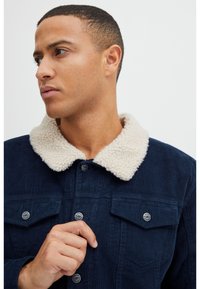Chaqueta de pana navy con un cuello de peluche crema, que presenta botones, dos bolsillos en el pecho y un tejido texturizado. Primer plano de la tela y los detalles.