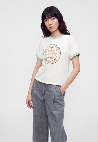 Femme aux cheveux noirs portant un T-shirt blanc avec bordure verte et texte graphique orange, porté avec un pantalon gris ample, debout avec une main dans la poche.