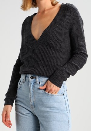 Femme portant un pull côtelé gris foncé à col en V avec manches longues, rentré dans un jean bleu clair taille haute, main dans la poche.