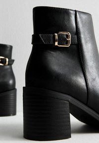 Botas negras hasta el tobillo de cuero con una superficie texturizada. Incluyen un adorno de hebilla lateral y un tacón grueso y estriado.