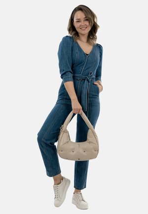 Giovane donna in un tuta di denim blu che tiene una borsa beige con borchie, indossando sneakers bianche, sorridendo su uno sfondo chiaro.