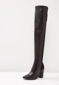 Bota negra por encima de la rodilla hecha de cuero brillante, con punta afilada, tacón grueso y detalles de tachuelas a lo largo de la suela. Textura suave.