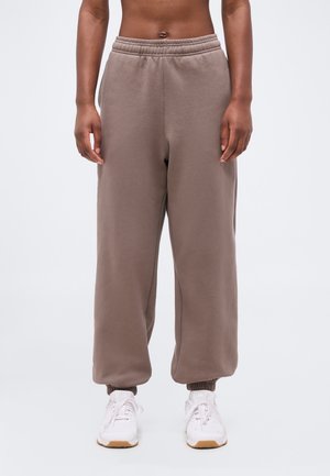 Άτομο που φοράει χαλαρά καφέ παντελόνια φόρμας με ελαστικά τελειώματα και λευκά αθλητικά παπούτσια, στέκεται μπροστά σε απλό ανοιχτό φόντο.