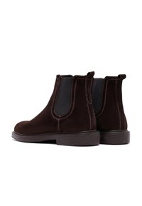 Donkerbruine suède Chelsea-boots met elastische zijpanelen, trekflappen aan de achterkant en lage gestapelde zolen, weergegeven tegen een witte achtergrond.