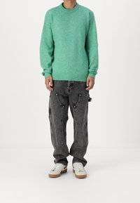 Zelený pletený svetr s kulatým výstřihem ve spojení s nadměrnými šedými riflovými kalhotami s kapsami a vybledlou texturou. Dokončeno bílými teniskami.