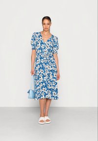 Vestido midi floral azul com mangas balão, decote em v e cintura ajustada. Combinado com sandálias brancas e uma bolsa azul clara.