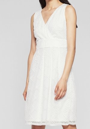 Robe blanche sans manches longueur genou avec encolure en V et motif géométrique texturé portée par une femme debout sur un fond uni.