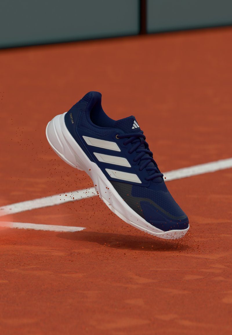 Chaussure de sport bleu marine avec des accents blancs, tige en mesh, semelle texturée et trois bandes blanches, suspendue au-dessus d'une surface de court en terre battue.