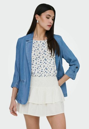 Femme aux longs cheveux bruns, portant des boucles d'oreilles argentées, un blazer bleu, un haut blanc à motifs floraux, et une jupe blanche à volants, se tenant avec une main dans la poche.