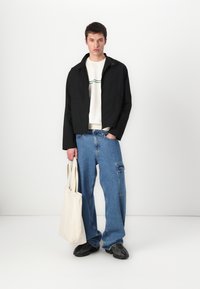 Jachetă neagră cu guler înalt, hanorac alb cu dungi verzi, pantaloni cargo din denim albastru lejer și un rucsac bej.