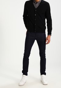 Svart cardigan med V-hals och knappar, bärs över en grå skjorta, matchad med mörka jeans och vita sneakers, står på en ljus bakgrund.