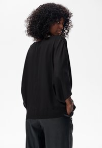 Schwarze, locker sitzende Bluse mit Dreiviertelärmeln, glatter Textur, subtiler Drapierung und rundem Ausschnitt. Elegantes und einfaches Design.