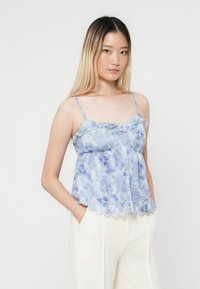 Lichtblauwe bloemenhemd met ruffles aan de rand en knoopdetails, gemaakt van een doorschijnende stof. Model draagt een crèmekleurige broek met wijde pijpen.