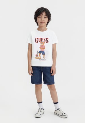Ragazzo con t-shirt bianca con stampa di orso e barca, pantaloncini blu navy, calzini bianchi e sneakers bianche in piedi contro uno sfondo semplice.