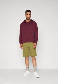 Hoodie com fecho de correr na cor bordô com detalhes pretos, combinado com calções verde-oliva. Ténis brancos e meias cinzentas com logótipo completam o conjunto.