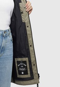 Vandafvisende jakke med sort ydre og en kontrasterende olivenfarvet foring. Indeholder et stort logo-patch og knaplukninger. Letvægtsmateriale.