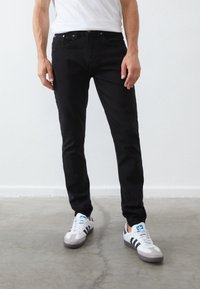 Homme portant un jean noir slim, un t-shirt blanc et des baskets Adidas blanches avec des bandes noires, debout sur un sol en béton.