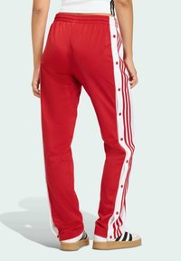 Červené športové nohavice s bielymi bočnými pruhmi, cvokmi a elastickým pásom. Vyrobené z hladkej tkaniny, so strihom rovného nohu.