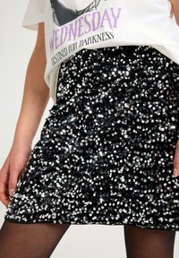 Nærbillede af en person iført en hvid "Wednesday Destined for Darkness" t-shirt og en sort palietteret miniskørt med sorte strømpebukser.