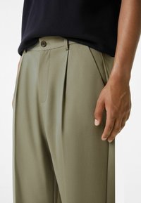 Světlé olivově zelené kalhoty s bočními záhyby, plochým předním dílem a zapínáním na knoflík. Tkanina má hladkou, strukturovanou texturu.