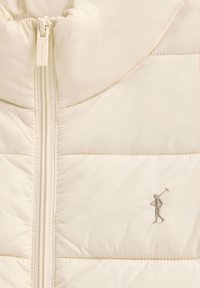 Närbild på beige quiltad jacka med främre dragkedja och liten broderad logotyp av en polospelare på bröstet.