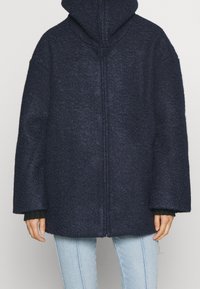 Navyblå hooded kappa i texturerat tyg, med dragkedja, vida ärmar och avslappnad passform. Ljusblå jeans syns under.