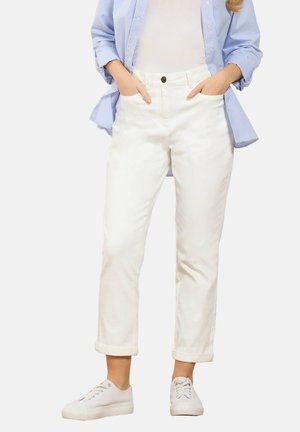 Kvinde iført opkrogede hvide bukser, hvide sneakers, en hvid top og en blåstribet skjorte med knapper, med hænderne i lommerne.