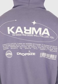 Lila Kapuzenpullover mit weißem Text "KARMA" und dem Spruch "Tu Gutes und Gutes wird dir folgen", sowie kleinerem motivierendem Text und Markenzeichen.