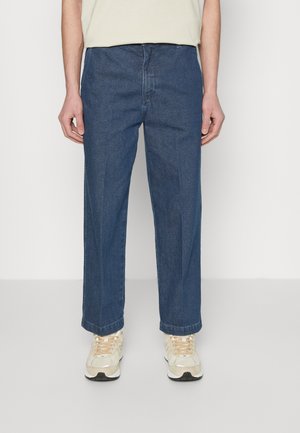 Mand iført blå denimjeans med lige ben og beige sneakers, stående mod en ensfarvet, lys baggrund.