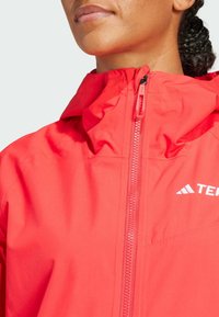 Red waterproof jacket with a high collar, seamless zippers, and white logo branding. Smooth texture and lightweight design for outdoor use.