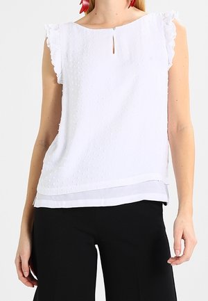 Blouse blanche à manches courtes avec des pois texturés, décolleté à ouverture, manches froncées et ourlet superposé, accompagnée de pantalons noirs.
