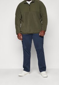 Uomo che indossa un pullover in pile verde oliva, pantaloni blu navy e sneakers bianche, in piedi contro uno sfondo neutro.