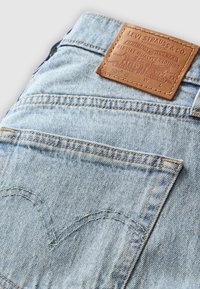 Poche arrière de jean en denim bleu clair avec surpiqûres jaunes et écusson en cuir marron de la marque Levi Strauss & Co. au-dessus de la poche.