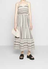 Femme portant une robe maxi sans manches à motifs noir et blanc, tenant une pochette en blanc texturé et portant des sandales à plateforme noires.