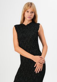 Vrouw met blond haar in een mouwloze zwarte textuurjurk, staand met de handen voorzichtig voor zich gekruist tegen een effen witte achtergrond.