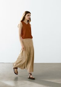Top senza maniche a V in rust abbinato a una maxi gonna plissettata beige. Texture morbide, design minimalista e sandali con cinturini marroni completano il look.