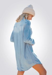 Vestido camisero de denim azul claro con mangas largas, puños abotonados y bolsillos laterales. Presenta un dobladillo curvado y detalle de yugo en la parte trasera.