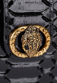 Czarna, teksturowana skóra z klamrą w kolorze złotym i czarnym, ozdobioną klejnotami, w kształcie stylizowanej głowy gada.