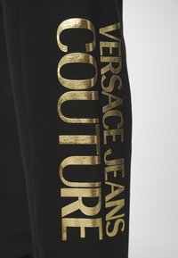Schwarze Jogginghosen mit großen, glänzenden goldenen Schriftzügen "VERSACE JEANS COUTURE". Weicher Stoff mit einfachem, figurbetontem Design.