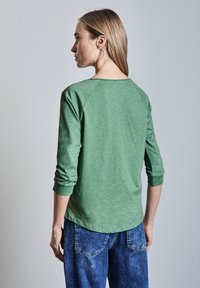 Vihreä pitkähihainen paita, jossa on rento leikkaus ja raglan-hihat, valmistettu pehmeästä kankaasta. Yhdistetty vaaleansinisiin farkkuihin.