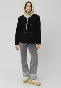 Veste en polaire noire avec de grands boutons toggle, associée à un jean flare gris et des chaussures à plateforme beiges. Le mannequin porte une légère capuche.