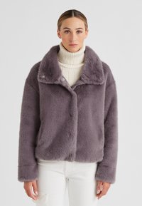 Lila Kunstfelljacke mit breitem Kragen, Knopfverschluss und langen Ärmeln, getragen über einem cremefarbenen Rollkragenpullover. Einfarbiger Hintergrund.