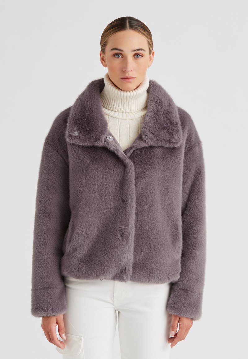 Lila Kunstfelljacke mit breitem Kragen, Knopfverschluss und langen Ärmeln, getragen über einem cremefarbenen Rollkragenpullover. Einfarbiger Hintergrund.