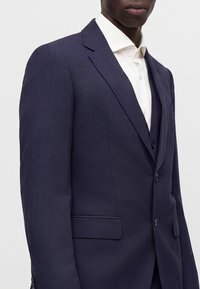Homme portant une veste de costume bleu marine sur une chemise blanche, visible des épaules jusqu'au milieu du torse sur un fond uni.