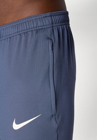 Nærbilde av blå Nike treningsshorts med elastisk midje og glidelås-lomma, båret av en person med mørk hud.
