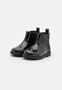 Botas de tobillo de charol negro con un acabado brillante, que presentan un acento de purpurina negra texturizada en el tacón y una lengüeta para facilitar el uso.