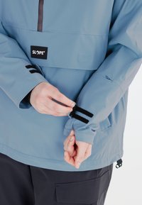 Helle blaue wasserdichte Jacke mit einem Reißverschluss auf der Vorderseite, verstellbaren Bündchen mit schwarzen Klettverschlussriemen und einem SLOPE-Logo auf der Brust.